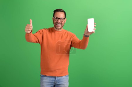 Joven vendedor alegre mostrando el cartel de pulgares hacia arriba y la pantalla del teléfono inteligente en un fondo verde aislado