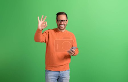 Retrato del hombre sonriente positivo usando un teléfono inteligente y mostrando el signo OK aislado sobre un fondo verde