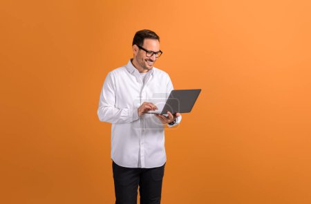 Retrato de sonriente hombre de negocios confiado haciendo investigación en línea sobre el ordenador portátil en fondo naranja