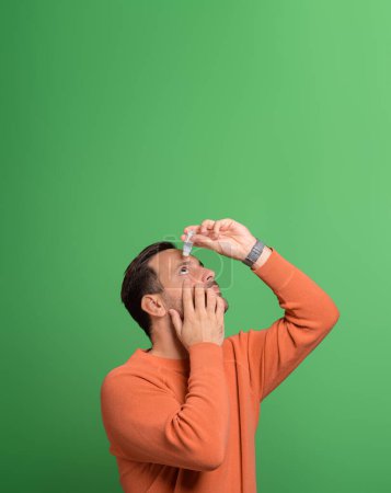 Portrait of overworked young man using eyedropper for treating irritated eyes on green background