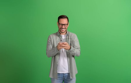 Excited young man in eyeglasses chatting online over mobile phone against isolated green background