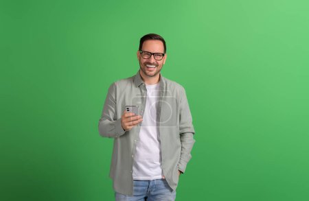 Porträt eines gutaussehenden Unternehmers mit Brille, der auf grünem Hintergrund lächelt und sein Handy benutzt