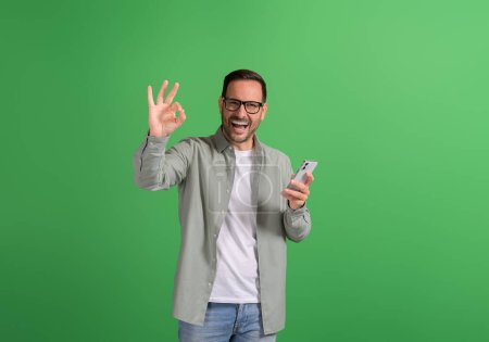 Junger Mann lacht fröhlich und zeigt OK-Geste, während er Handy auf grünem Hintergrund benutzt