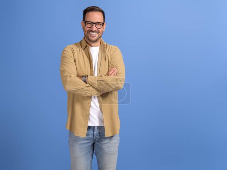 Happy young businessman with eyeglasses and arms crossed posing confidently on blue background