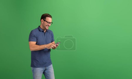 Retrato de un joven confiado sonriendo y revisando correos electrónicos sobre un teléfono inteligente sobre un fondo verde