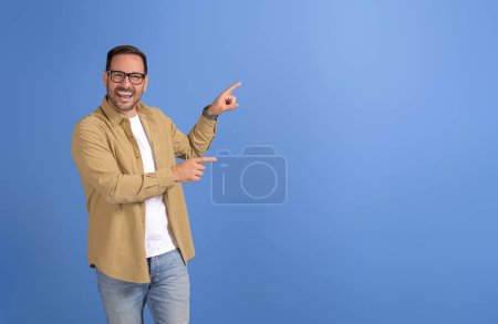 Porträt eines jungen Verkäufers, der lachend auf eine leere Werbefläche auf blauem Hintergrund zielt