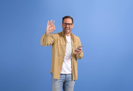Porträt eines fröhlichen jungen Mannes, der per Handy Nachrichten sendet und OK-Geste auf blauem Hintergrund zeigt