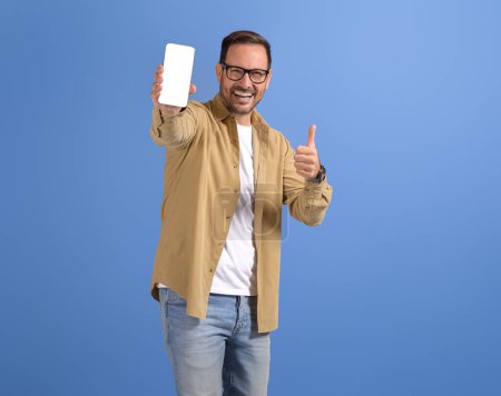 Porträt eines glücklichen jungen Mannes, der den leeren Bildschirm des Smartphones zeigt und das Zeichen "Daumen hoch" auf blauem Hintergrund zeigt