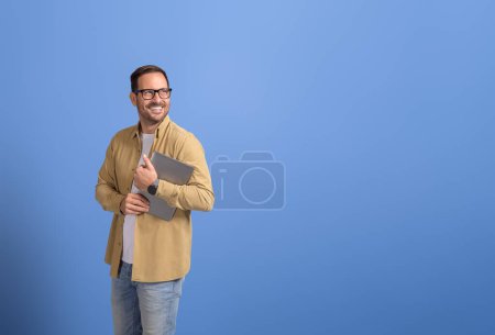 Portrait of positive young businessman holding laptop and looking away cheerfully on blue background