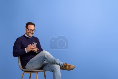 Porträt eines lächelnden Geschäftsmannes, der auf einem Stuhl sitzt und vor blauem Hintergrund über sein Smartphone SMS schreibt