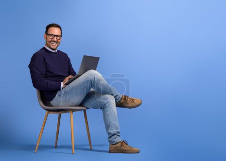 Portrait d'homme souriant professionnel et utilisant un ordinateur sans fil sur chaise sur fond bleu