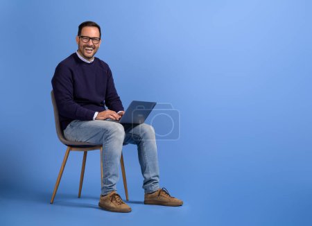 Portrait de gai manager masculin marketing en ligne sur ordinateur sur chaise sur fond bleu