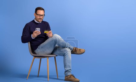 Geschäftsmann kauft online mit Handy und Kreditkarte ein, während er auf einem Stuhl auf blauem Hintergrund sitzt
