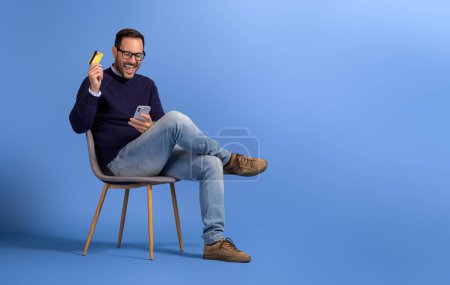 Aufgeregter junger Mann mit Kreditkarte und Handy kreischt fröhlich auf Stuhl vor blauem Hintergrund
