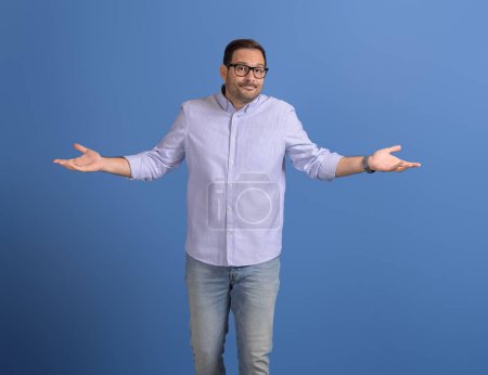 Confused businessman spreading hands and shrugging shoulders while standing against blue background