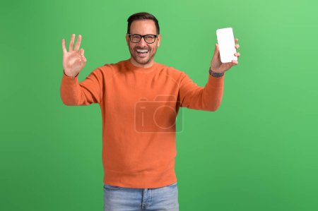 Portrait of happy young businessman showing mobile phone screen and OK sign against green background