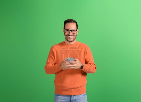 Handsome smiling businessman in eyeglasses messaging over smart phone on isolated green background