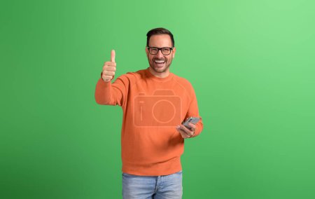 Retrato de joven empresario sonriente mostrando como signo y utilizando el teléfono móvil sobre fondo verde