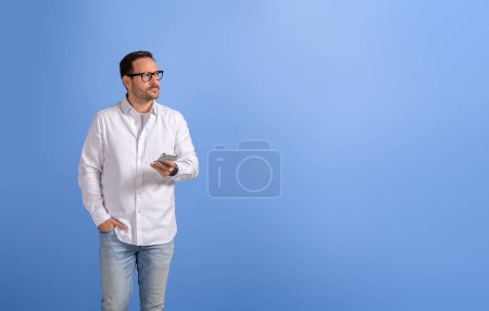 Hombre de negocios serio sosteniendo el teléfono móvil y las ideas de pensamiento mientras mira hacia otro lado en el fondo azul