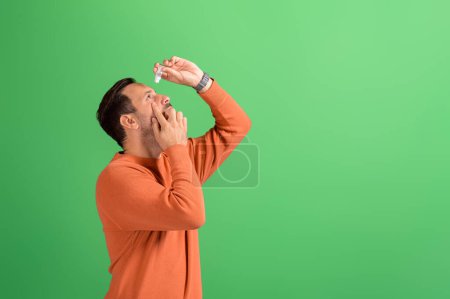 Side view of young man with conjunctivitis putting drops for treatment on isolated green background