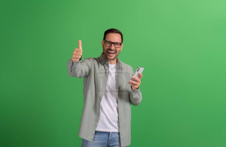 Portrait of ecstatic young salesman showing thumbs sign and using mobile phone on green background