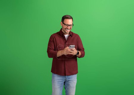 Hombre joven guapo en gafas sonriendo y mensajes de texto en línea sobre el teléfono inteligente sobre fondo verde