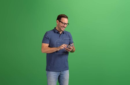 Handsome young man in glasses smiling and checking messages over mobile phone on green background