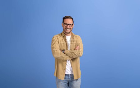Portrait of handsome young manager with arms crossed and in eyeglasses smiling over blue background