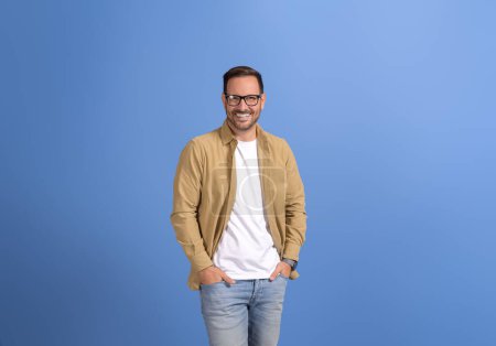 Portrait of handsome young man in eyeglasses and with hands in pockets standing on blue background