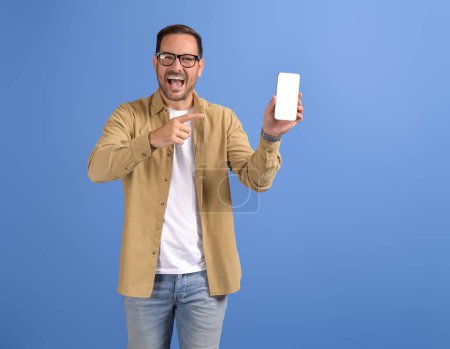 Porträt eines fröhlichen jungen Mannes, der auf Smartphone und Werbe-Dating-App auf blauem Hintergrund zeigt