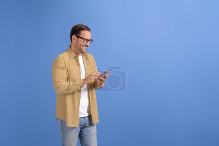 Happy young businessman in glasses smiling and texting online over smart phone on blue background
