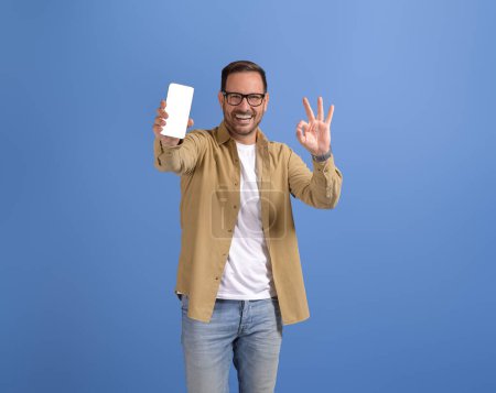 Feliz vendedor mostrando signo OK y publicidad aplicación de compras en línea sobre el teléfono en fondo azul