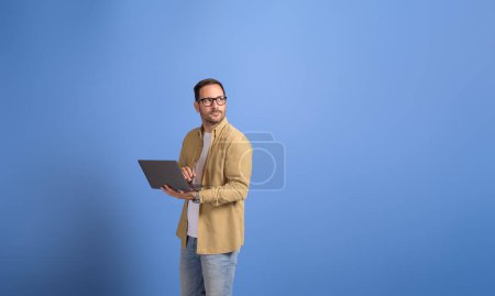 Seriöser junger Geschäftsmann hält Laptop in der Hand und blickt nachdenklich über isolierten blauen Hintergrund hinweg