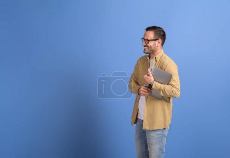 Retrato de un joven empresario sonriente sosteniendo una computadora portátil y mirando hacia otro lado sobre un fondo azul