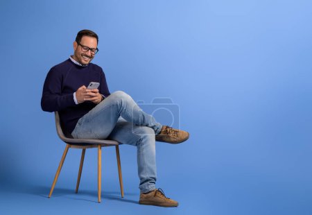 Schöner junger Mann lächelt und nutzt soziale Medien per Smartphone auf Stuhl vor blauem Hintergrund
