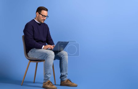 Portrait d'entrepreneur masculin lisant des e-mails sur ordinateur portable et assis sur une chaise sur fond bleu