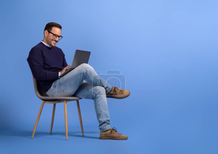 Téléchargez les photos : Confiant jeune homme d'affaires dans les lunettes à l'aide d'un ordinateur portable tout en étant assis sur une chaise sur fond bleu - en image libre de droit