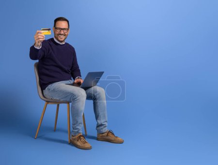 Glücklicher männlicher Kunde zeigt Kreditkarte und Einkauf über Laptop auf Stuhl vor blauem Hintergrund