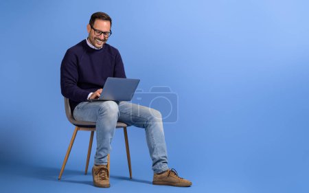 Heureux freelance masculin travaillant sur le projet sur ordinateur portable tout en étant assis sur la chaise sur fond bleu