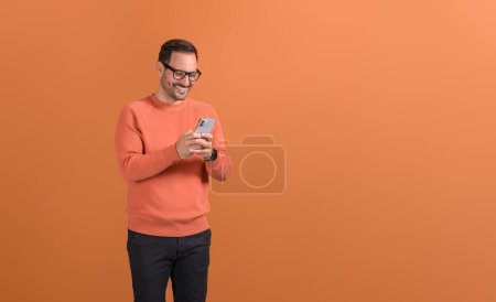 Freelancer masculino sonriente revisando aplicaciones de redes sociales a través del teléfono móvil sobre fondo naranja