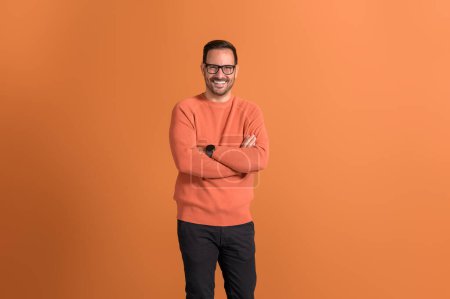 Portrait of young happy businessman in eyeglasses smiling with arms crossed on orange background