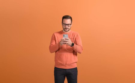 Serious young man in eyeglasses texting online over smart phone while standing on orange background