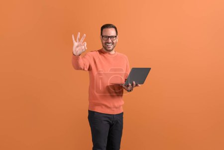 Gut gelaunte Freiberuflerin mit Laptop zeigt selbstbewusst OK-Zeichen auf orangefarbenem Hintergrund