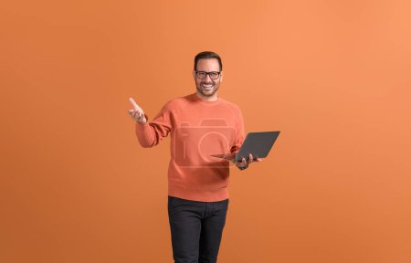 Handsome entrepreneur holding laptop and gesturing confidently while standing on orange background