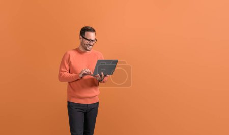 Souriant beau pigiste travaillant en ligne sur un ordinateur sans fil sur fond orange isolé
