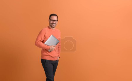 Beau entrepreneur avec la main dans la poche tenant ordinateur portable et souriant à la caméra sur fond orange