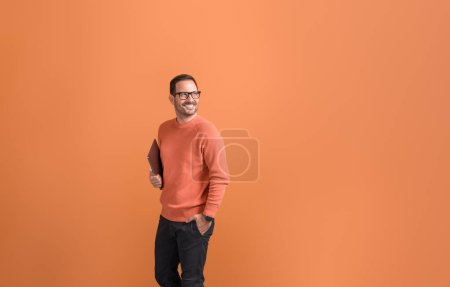 Contemplativo joven empresario sosteniendo el ordenador portátil y mirando hacia otro lado mientras está de pie sobre fondo naranja