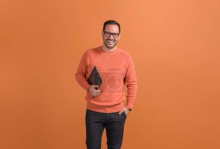 Réussi homme freelance heureux avec la main dans la poche tenant ordinateur portable et posant sur fond orange