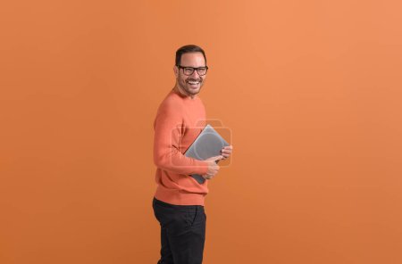 Portrait of cheerful young man holding laptop and laughing at camera on isolated orange background
