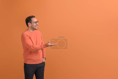 Jungunternehmer lacht und wirbt auf leerer Handfläche vor orangefarbenem Hintergrund für ein neues Produkt